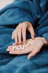 Woman sitting on bed wrapped in blue blanket, holding pills in hands