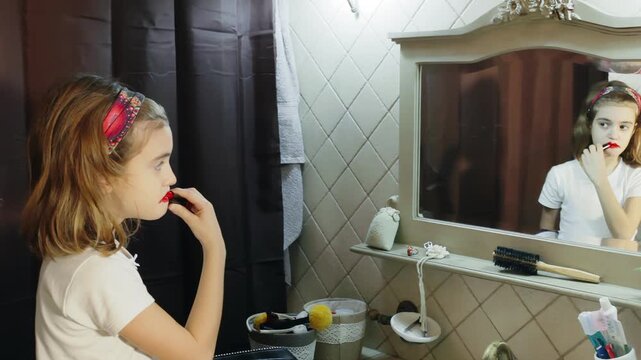Young girl applies red lipstick in bathroom mirror, smearing it across her cheeks. Her expression shows curiosity and excitement as she explores makeup, creating an endearing and humorous scene.
