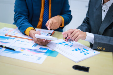 Two businessmen are seated at a desk in an office, engaged in a meeting focused on analysis, finance, and marketing strategies, with documents and graphs spread across the table.