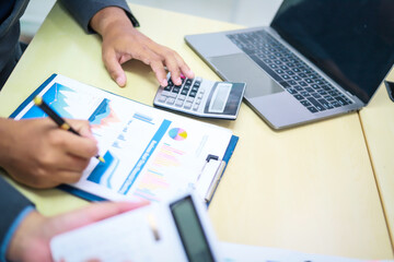 Two businessmen are seated at a desk in an office, engaged in a meeting focused on analysis, finance, and marketing strategies, with documents and graphs spread across the table.