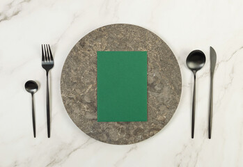 Top view of brown marble plate, black cutlery and green card on white marble background. Food...