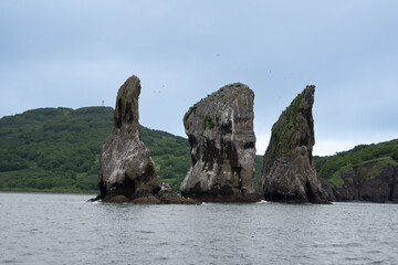 From the sea side there are Three brothers rocks in the Avacha Bay. In the background there are green mountains and hills.