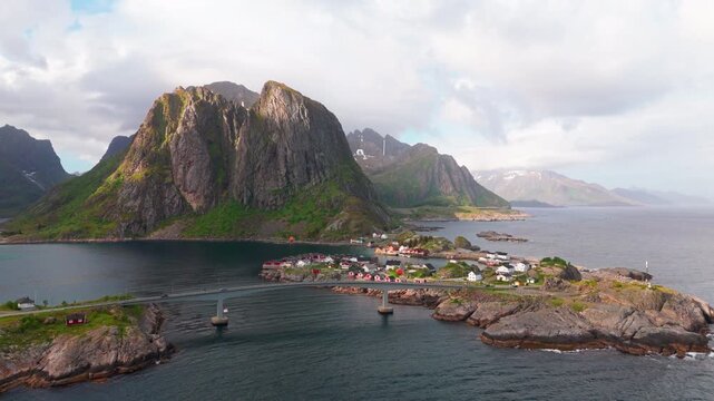 Camera asacending, Hamnoy village in the foreground and Lilandstinden mountain peak in the background. Lofoten, Norway. Red fisherman's cabin on small islands, cars crossing archipelago bridges.
