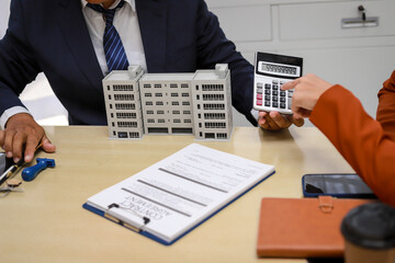 A mid-adult businessman in a modern office is seated at a desk, reviewing documents with a client. They discuss real estate investments and home insurance. A calculator and laptop are nearby