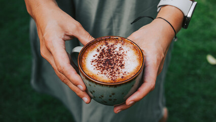 Female hands holding ceramic mug with cappuccino on a green background. Concept of delicious cinnamon coffee close-up