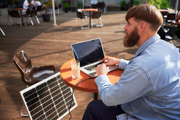 Man works on laptop at outdoor round table, with glass of iced coffee. Solar panel nearby,...