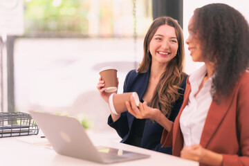 Two young businesswomen work together on laptops while talking about panning, reading documents and writing notes while discussing a new project.