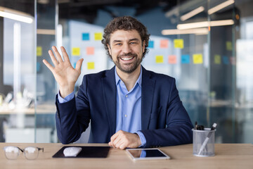 Business professional waving at the camera during a video call in an office. He smiles confidently, surrounded by a modern work environment with colorful sticky notes on glass walls.