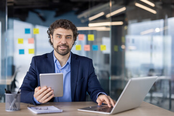 A confident businessman in a modern office using a tablet and laptop, showcasing digital communication and work productivity.