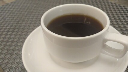 A close-up shot of a white ceramic cup filled with hot black coffee placed on a matching saucer. The cup is positioned on a textured table, perfect for use in food and beverage promotions.