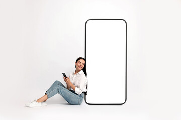 Beautiful young woman with smartphone sitting near big mobile phone with blank screen on light...
