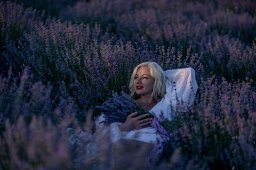 Blonde woman poses in lavender field at sunset. Happy woman in white dress holds lavender bouquet. Aromatherapy concept, lavender oil, photo session in lavender