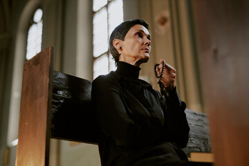 Senior woman dressed in total black sitting on wooden bench and holding wooden rosary in church