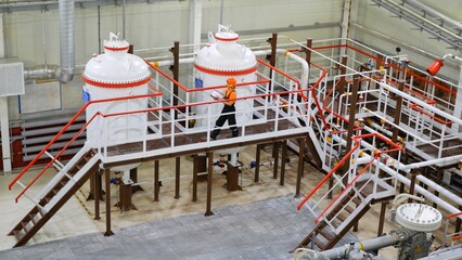 Engineer wearing orange vest and helmet walks around water treatment tanks and equipment on metal...