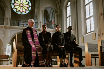 Senior pastor and sorrowful relatives dressed in total black sitting on bench during funeral in...