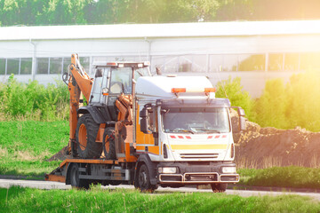 Orange tow truck hauls excavator on flatbed trailer down rural road in summer