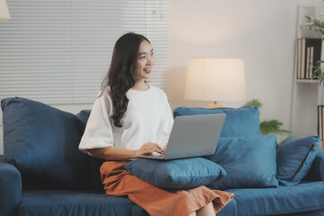 Young asian freelancer is working from her home office, sitting comfortably on a blue sofa using a laptop computer while looking away from the screen with a cheerful smile