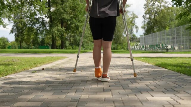 teenage girl walks gingerly forward on crutches through a summer park, her leg in a cast. View from behind.