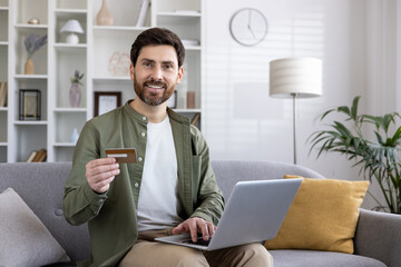 Confident man sitting on sofa holding credit card and using laptop for online shopping. Shows convenience and enjoyment of digital transactions at home. Ideal for themes of modern lifestyle