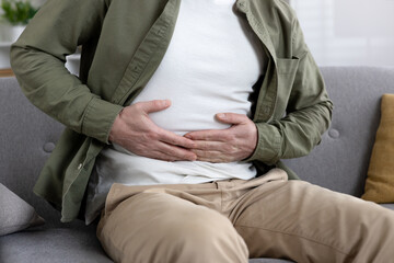 Man sitting on couch experiencing abdominal pain holding stomach. Wearing casual clothing and showing visible discomfort. Concept of stomachache, digestive issues, or health concerns.