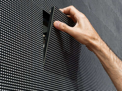 A man's hand removing a square LED module from its place on the surface of a large LED screen for repair and maintenance