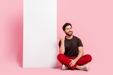 Full size photo of nice man dressed brown t-shirt red pants sit indicating at white wall empty space isolated on pink color background