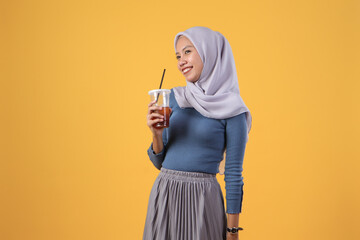 portrait of happy asian indonesian muslim woman drinking cold ice tea in plastic cup on isolated yellow background