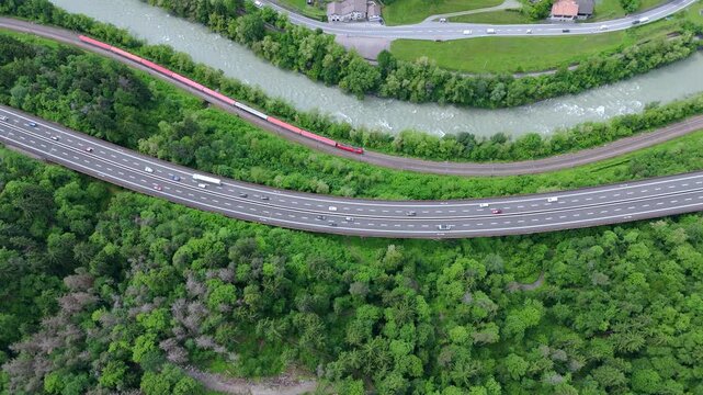 Aerial footage of a highway and parallel train tracks curving through a dense, green forest with a river flowing nearby.