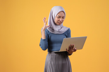 asian indonesian muslim woman holding laptop computer with angry face expression on isolated yellow background