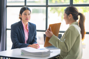 Two Asian businesswomen use laptop and tablet to collaborate in a productive conversation with colleagues while analyzing documents, financial graphs, working in the office.