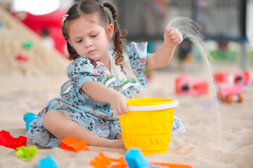 A little girl is playing in the sand with a yellow bucket. She is wearing a blue dress and has her hair in pigtails