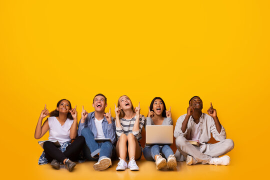 Excited multiracial students pointing up at copy space, sitting on floor over yellow background, big discounts for students