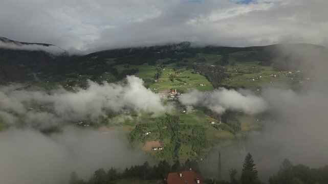 Aerial view of Villanders village enveloped in morning clouds, surrounded by lush green fields and forested hills in a picturesque mountain valley.