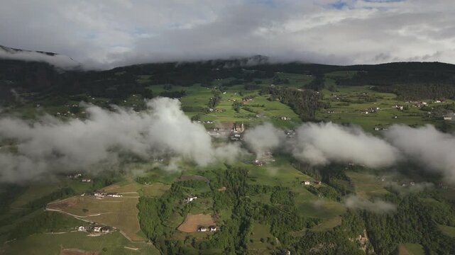 Aerial footage of a mountain valley with patches of clouds drifting over green fields and small villages, surrounded by forested hills.