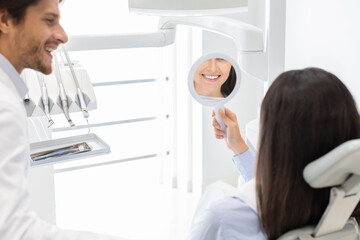 Obraz premium Sattisfied young woman client of dental clinic smiling at mirror, handsome dentist standing next by