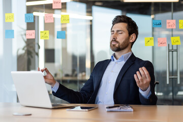 Businessman practicing meditation at desk surrounded by sticky notes and laptop. Focus on stress relief, mindfulness, and mental clarity in busy office environment. Relaxation techniques in workplace