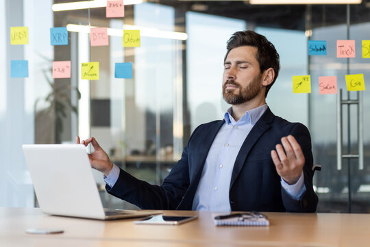 Businessman practicing meditation at desk surrounded by sticky notes and laptop. Focus on stress relief, mindfulness, and mental clarity in busy office environment. Relaxation techniques in workplace