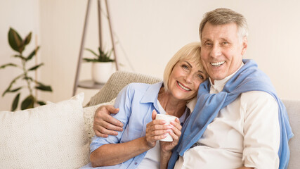 Happy senior couple relaxing on sofa and enjoying morning tea, man showing thumb up