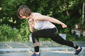 Fototapeta premium An adult woman is doing fitness in the park. The woman is doing lunges and squats.
