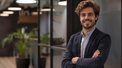 Fototapeta premium Smiling handsome businessman with crossed arms in modern office, young man exuding confidence, business success concept.