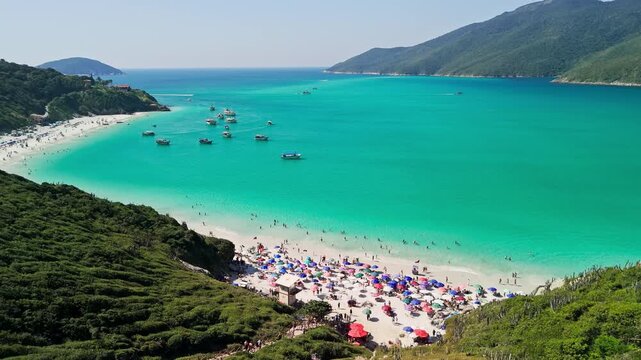Drone rises, panning left, revealing white sandy beaches, turquoise waters, anchored boats, and majestic island mountains in Arraial do Cabo