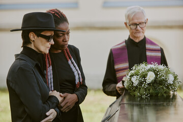 Gray-haired pastor reading prayer while mother and daughter bemoaning their loss