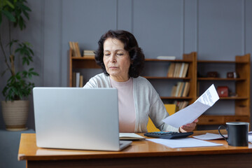 Middle aged senior woman sit with laptop and paper document. Pensive older mature lady reading paper bill pay online at home managing bank finances calculating taxes planning loan debt pension payment