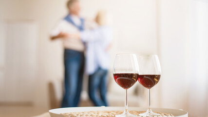 Celebrate anniversary. Mature couple in love dancing on background, glasses with red wine on foreground