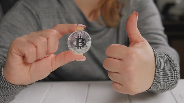 A close-up of a silver bitcoin coin in the hands of a young business woman. The woman gives a thumbs up. The concept of finance and investment, cryptocurrency and bitcoin trading.