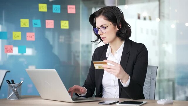 Businesswoman holding credit card entering data on a laptop in office with shocked expression, realizing she is a victim of fraud and deception. Concept of online scam, financial crisis, cybersecurity