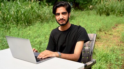 Indian young boy doing work from home. Man sitting on office table at garden doing work on internet