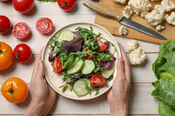Healthy vegetarian food. Woman holding plate of salad at light wooden table with vegetables, top view