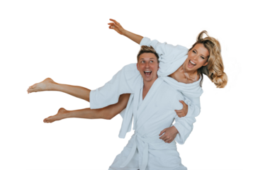 Playful couple in bathrobes having fun on a hotel bed, with the man carrying the woman on his back, both laughing and enjoying a joyful moment together