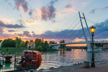 Latarnia na promenadzie na tle mostu i zachodzącego słońca w Mikołajkach   A lantern on the promenade on the bridge and the setting sun background in Mikołajki Town © Adrian White